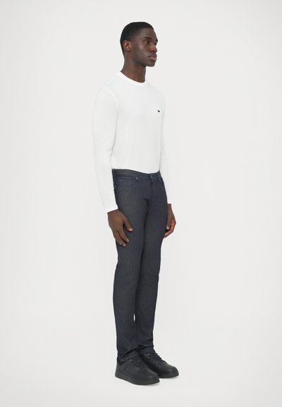 Λευκό πουκάμισο με μακριά μανίκια και στρογγυλή λαιμόκοψη, συνδυασμένο με σκο�ύρο μπλε τζιν. Μαύρα αθλητικά παπούτσια ολοκληρώνουν το σύνολο. Το μοντέλο στέκεται σε προφίλ.