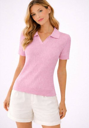 Femme portant un haut en maille rose à manches courtes avec un col et un short blanc taille haute, debout devant un fond uni clair.