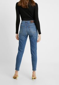 Jeans de mezclilla azul de cintura alta en un corte ajustado, con bolsillos delanteros y traseros, y una etiqueta de cuero marrón en la cinturilla.