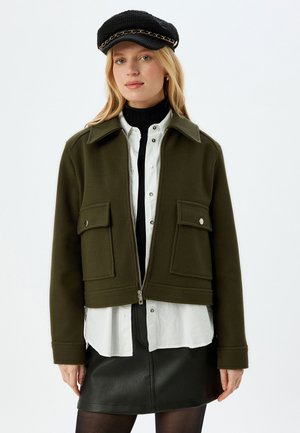 Olijfgroene spijkerjas met een ritssluiting aan de voorkant, twee grote klepzakken en een kraag. Gedragen over een witte blouse en een zwarte coltrui.