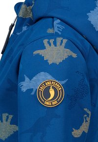 Blauwe jas met een mix van blauwe en gouden grafische patronen. Bevat een cirkelvormige 'Salt and Pepper' logo patch op de mouw.