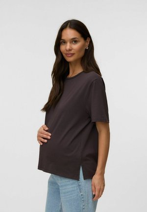 Femme enceinte aux longs cheveux foncés porte un t-shirt sombre et ample et un jean bleu clair, tenant doucement son ventre et souriant doucement.