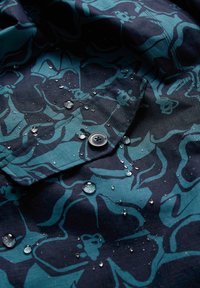 Blauw stoffen met bloemenpatroon en waterdruppels. Heeft een knoopdetail en een gestructureerd oppervlak, dat de waterdichte eigenschappen toont.