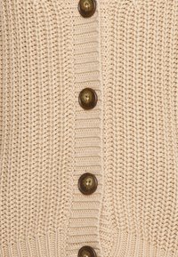 Béžová pletená tkanina s vertikálním žebrováním a čtyřmi hnědými kulatými knoflíky přišitými v jedné sloupci.