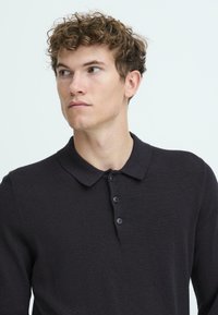 Sort strikking polo-shirt med krave, der har knaplukning og et tekstureret stof. Modellen har krøllet hår og en opretstående holdning.