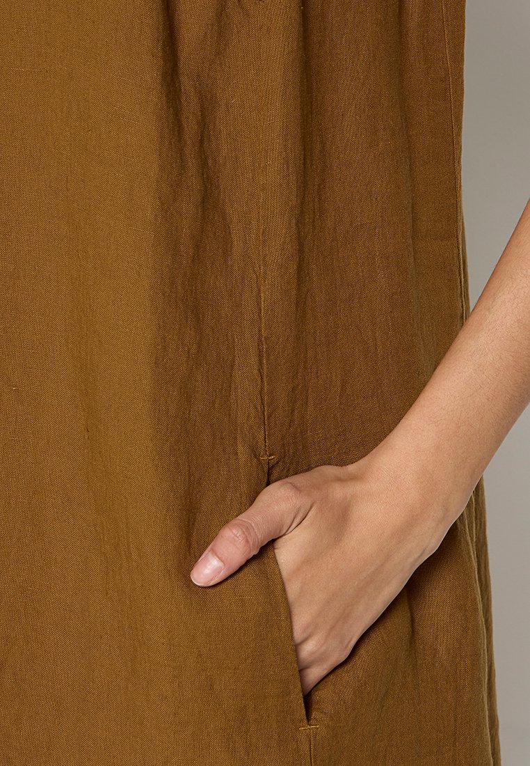 Brązowa lniana sukienka o teksturowanej powierzchni, z boczną kieszenią, w której umieszczona jest ręka, podkreślająca zarówno design, jak i funkcjonalność odzieży.