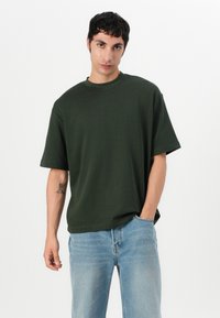 T-shirt en coton vert foncé avec une coupe loose, col rond et manches courtes, associée à un jean en denim bleu clair.