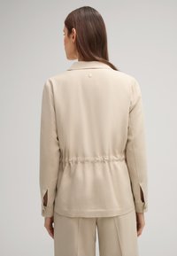 Giacca collare beige da donna, in tessuto leggero, con vita elastica, polsini con bottoni, dettagli posteriori sottili con piccolo accento circolare.