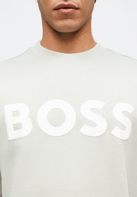 Άνδρας που φοράει γκρι ανοιχτό φούτερ με λαιμόκοψη, με μεγάλα λευκά γράμματα που σχηματίζουν τη λέξη «BOSS» στο στήθος.