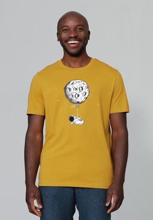 Geel katoenen T-shirt met een grafisch ontwerp van een astronaut die een ballon in de vorm van een maan vasthoudt, tegen een gedempte achtergrond.