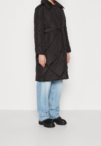 Osoba oblečená v černém prošívaném kabátu s páskem, světlých modrých kalhotách širokého střihu a černých masivních botách, stojí na jednoduchém bílém pozadí.