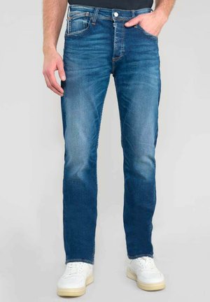 Jean en denim bleu avec une légère décoloration, coupe droite, design à cinq poches et coutures contrastantes, associés à des baskets blanches.