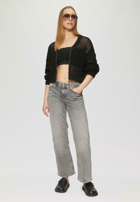 Schwarze Strickjacke mit offener Masche, schwarzes kurzes Oberteil, graue weite Jeans und schwarze Slipper. Das Model trägt oversized Sonnenbrillen.