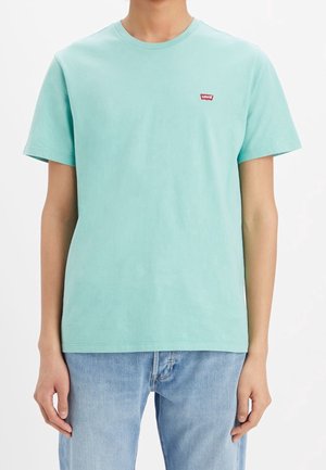 Persona che indossa una maglietta a maniche corte color turchese chiaro con collo rotondo e un piccolo logo rosso Levi's sul petto, abbinata a jeans azzurri.