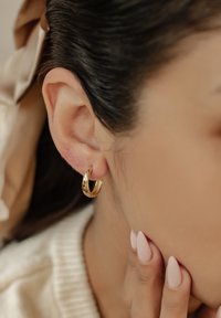 Créole en or accrochée à l'oreille inférieure, texture lisse, finition légèrement polie, forme circulaire. Fond doux et neutre avec des tons clairs.