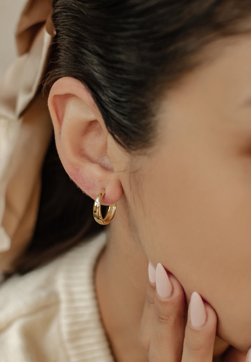 Créole en or accrochée à l'oreille inférieure, texture lisse, finition légèrement polie, forme circulaire. Fond doux et neutre avec des tons clairs.