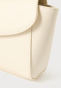 Gros plan sur un coin de sac à main en cuir beige montrant des coutures nettes et une fermeture à rabat sur fond blanc.