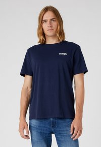 Camiseta de algodón azul marino con cuello redondo y mangas cortas; presenta un logo blanco de "Wrangler" en el lado izquierdo del pecho. Textura suave.