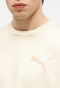 Κοντινό πλάνο ενός ατόμου που φοράει ένα εκρού πουκάμισο με ανυψωμένο λογότυπο Puma στο στήθος.