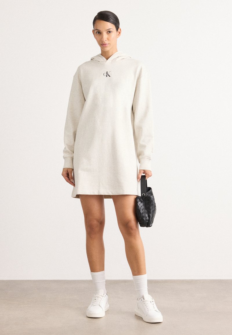 Beige hoodie jurk met een kangoeroe zak, ribbelmanchetten en een logo op de borst. Gepaard met witte sneakers en een zwarte geweven handtas.