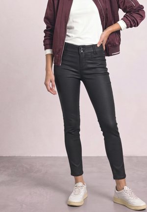 Femme portant un pantalon skinny noir, des baskets blanches, un haut blanc et une veste bomber bordeaux, debout avec une main sur la hanche.