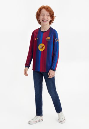 Dieťa s červenými vlasmi, ktoré má na sebe červeno-modrý pruhovaný dres FC Barcelona s dlhým rukávom, modré džínsy a biele tenisky, usmievajúce sa a stojace.