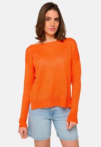 Femme se tenant debout, portant un pull orange à manches longues et un short en jean bleu clair, devant un fond blanc uni.