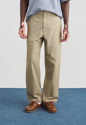 Beige Hose mit weitem Bein aus leichtem Stoff, mit einem Knopfverschluss und vorderen Taschen, kombiniert mit braunen Schuhen.