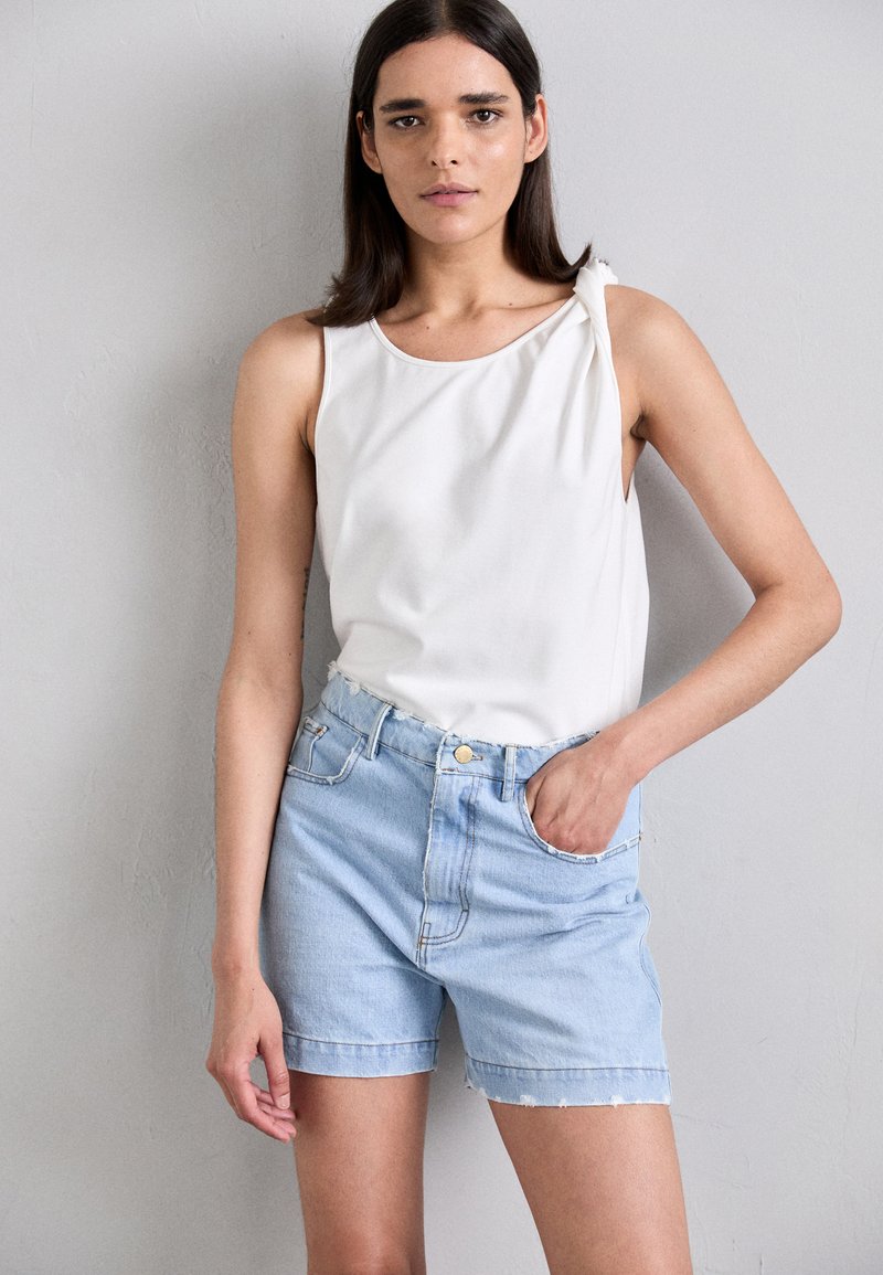Top bianco senza maniche con un nodo sulla spalla sinistra, abbinato a pantaloni corti in denim azzurro chiaro con orlo sfrangiato e tasche frontali.