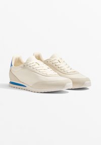 Beige Sneaker mit einem Obermaterial aus Wildleder und Leder, versehen mit einem blauen Akzent an der Ferse und einer weißen Gummisohle mit strukturiertem Muster.