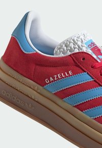 Červené semišové tenisky s modrými koženými pruhy, bílým síťovinovým jazykem a světlou gumovou podrážkou. Obsahují vyražené logo "Gazelle".