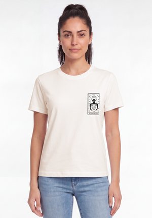 Femme portant un t-shirt blanc avec un dessin de carte de tarot noire intitulé Force montrant une femme et un lion sur la zone de la poche avant.