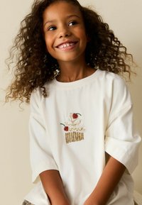 Valge suur üle suurusega t-särk, millel on tikitud koogikese kujundus värvikate helmeste ja litritega, punaste kirsimaitsete ja kuldsete aktsentidega.