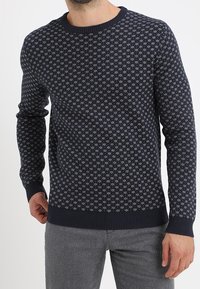 Navy blue knitted sweater featuring a geometric white pattern, round neck, and ribbed cuffs and hem. Warm and textured fabric.