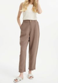 Pantalon en tissu léger marron avec une taille élastique, deux poches latérales et des boutons en bois en décoration. Porté avec un débardeur blanc et des sandales.