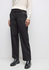 Femme portant un pantalon large à carreaux noirs, un pull beige et des bottines noires, debout devant un fond blanc uni.