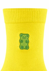 Gelbe gestrickte Socke mit einem gerippten Bund und einem grünen Gummibärchen, das an der Seite mit Punkten und Gesichtszügen gestickt ist.