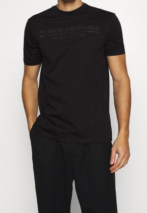 Mężczyzna ubrany w czarną koszulkę Armani Exchange z krótkim rękawem z ledwo widocznym logo i czarne spodnie, stoi z jedną ręką w kieszeni.