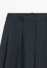 Pantaloni su misura grigio scuro con passanti per cintura e pieghe anteriori, mostrati dalla vita fino a metà coscia.
