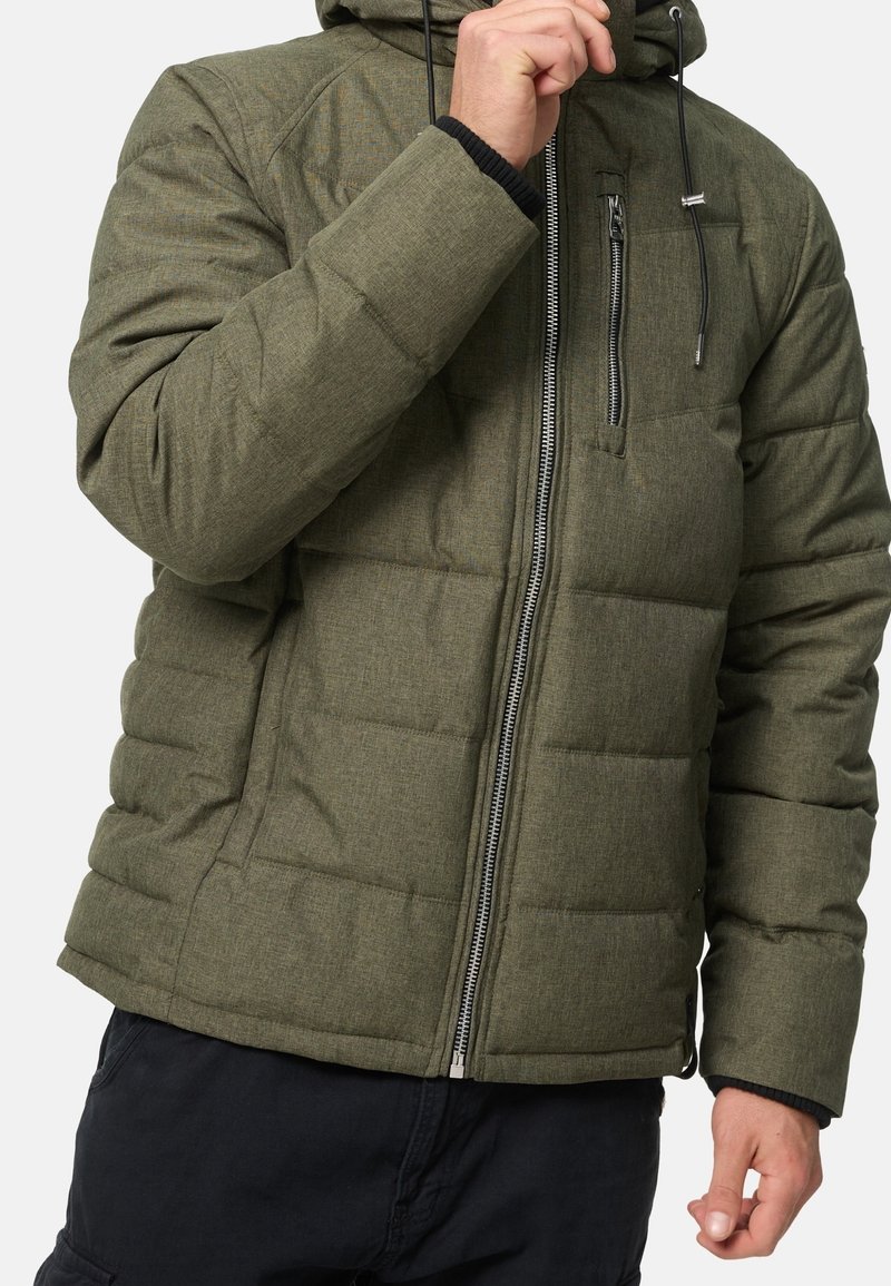 Personne portant une veste matelassée vert olive avec fermeture éclair frontale et poche poitrine, réglant le col de la veste d'une main.