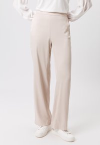 Pantalons beiges taille haute en tissu léger, avec une coupe droite. Associés à des baskets blanches, présentant une texture lisse.
