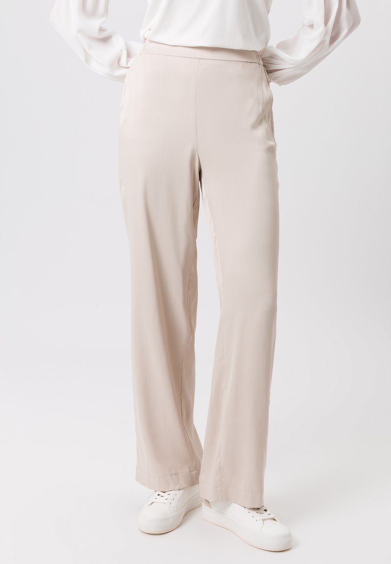 Pantalons beiges taille haute en tissu léger, avec une coupe droite. Associés à des baskets blanches, présentant une texture lisse.