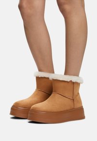 Botas de tobillo de ante marrón con forro de piel sintética blanca y una suela de plataforma de goma gruesa. Detalle de costura simple en la parte delantera.