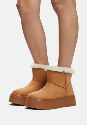 Tan suede ankle boots with white faux fur lining and a thick rubber platform sole. Simple stitched detail across the front.