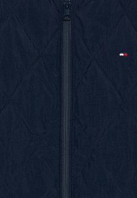 Marine quilted jakke med hel lengde glidelås, teksturert stoff, og lite logo med røde, hvite og blå detaljer på brystet.