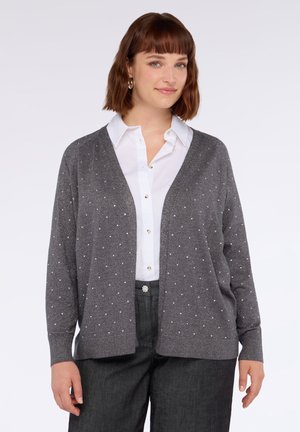 Junge Frau, die einen grauen Cardigan mit kleinen silbernen Nieten über einem weißen Hemd und dunkelgrauen Hosen trägt, steht vor einem schlichten Hintergrund.