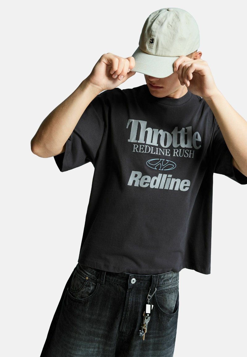 Črna prevelika majica s sivim grafičnim napisom "Throttle Redline Rush". Uporabljena z temno jeans hlačami, na katerih je ključno obroček na zanko pasu.