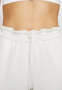 Bílý elastický pas s volánovými okraji, vybavený šňůrkovým uzávěrem. Hladká textura tkaniny s přiléhavým střihem.