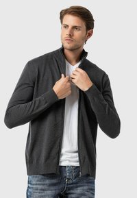 Mann trägt einen dunkelgrauen Reißverschluss-Pullover über einem weißen T-Shirt, richtet seinen Kragen und steht vor einem schlichten hellen Hintergrund.