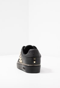 Sneaker nera Guess con borchie dorate e logo sul tallone e sulla suola, vista da dietro su una superficie bianca.